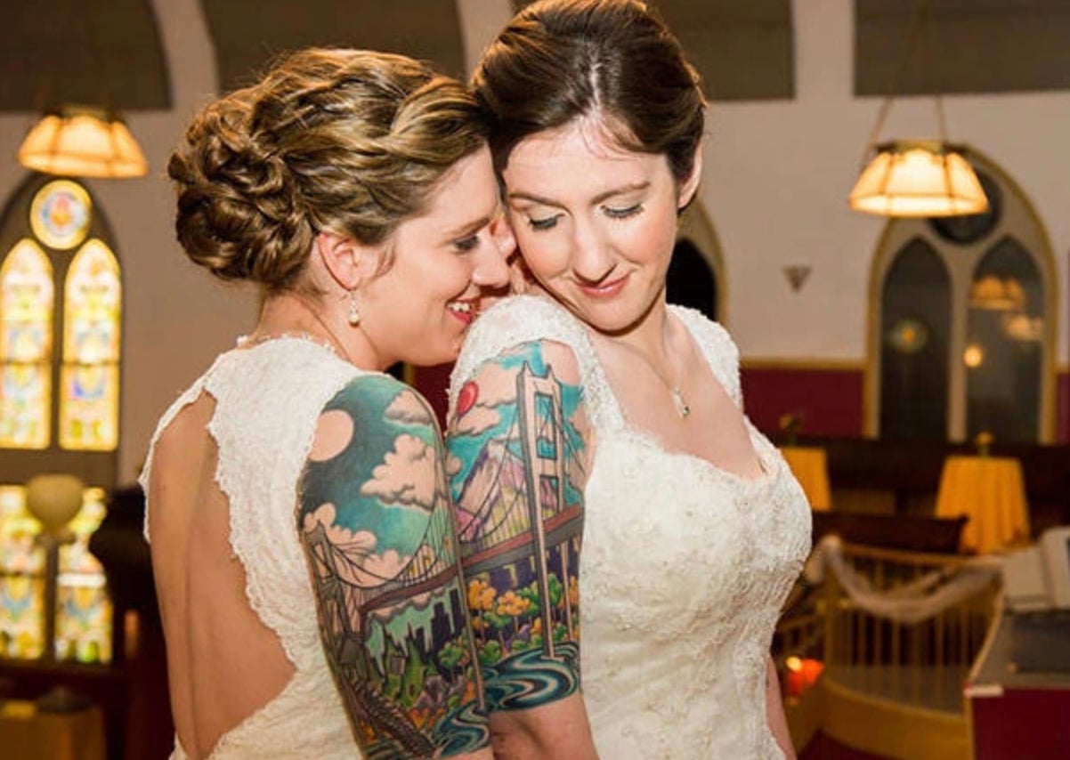 Two brides in white dresses stand close together in a warmly lit church. One bride smiles softly with eyes closed, while the other whispers with a smile, showing off a colorful bridge tattoo on her arm.