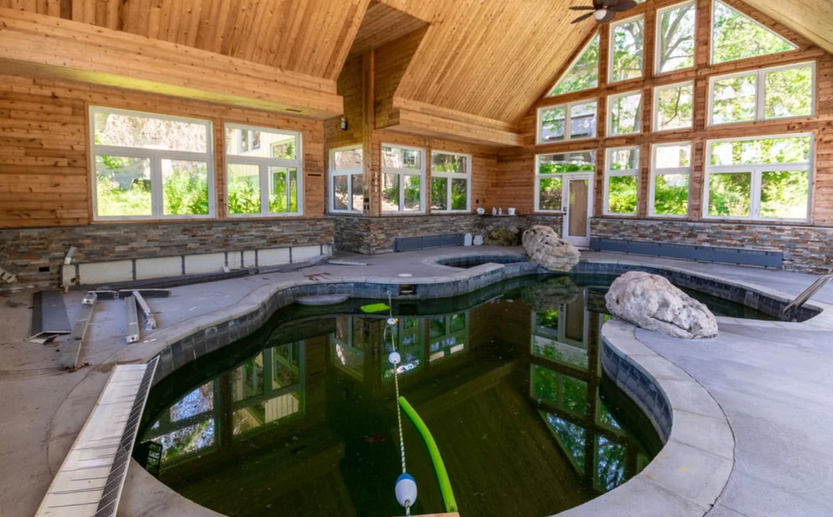 An indoor pool with a natural, irregular shape, surrounded by stone and wood-paneled walls. Large windows let in sunlight, and decorative boulders are placed around the poolside. The pool is partially uncovered.