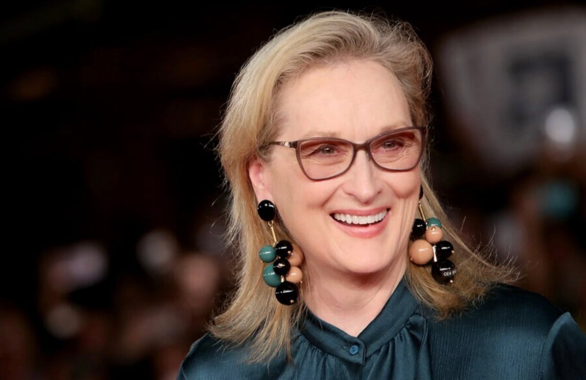 A smiling woman with blonde hair, wearing glasses, large statement earrings, and a dark green blouse, standing in front of a blurred background with bright lights.