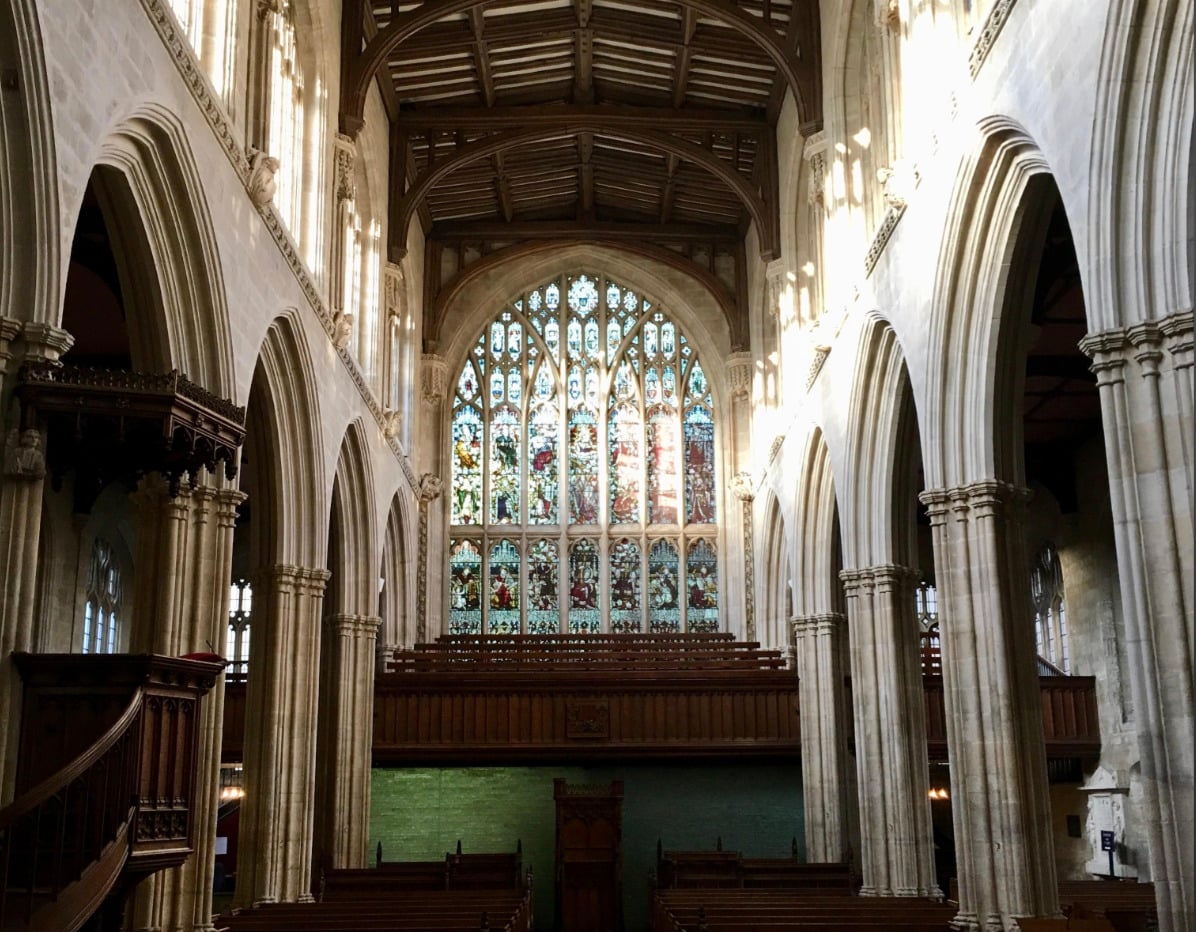 Interior of a historic church with tall stone arches, wooden pews, and a large, ornate stained glass window at the far end. Sunlight streams through the windows, illuminating the space.