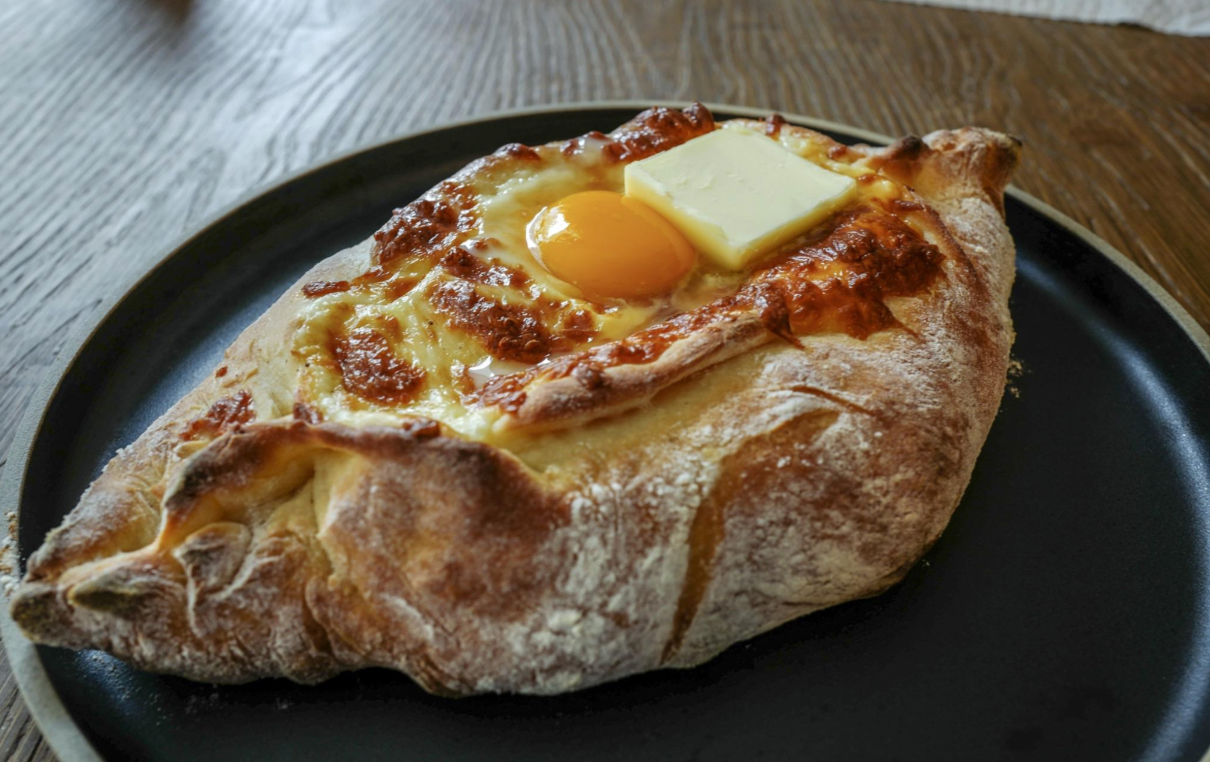 A khachapuri, a boat-shaped Georgian bread filled with melted cheese, a sunny-side-up egg, and a pat of butter, sits on a black plate atop a wooden table.