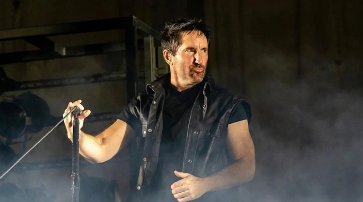 A man wearing a sleeveless black leather vest holds a microphone on stage, surrounded by smoke, with a serious expression on his face.