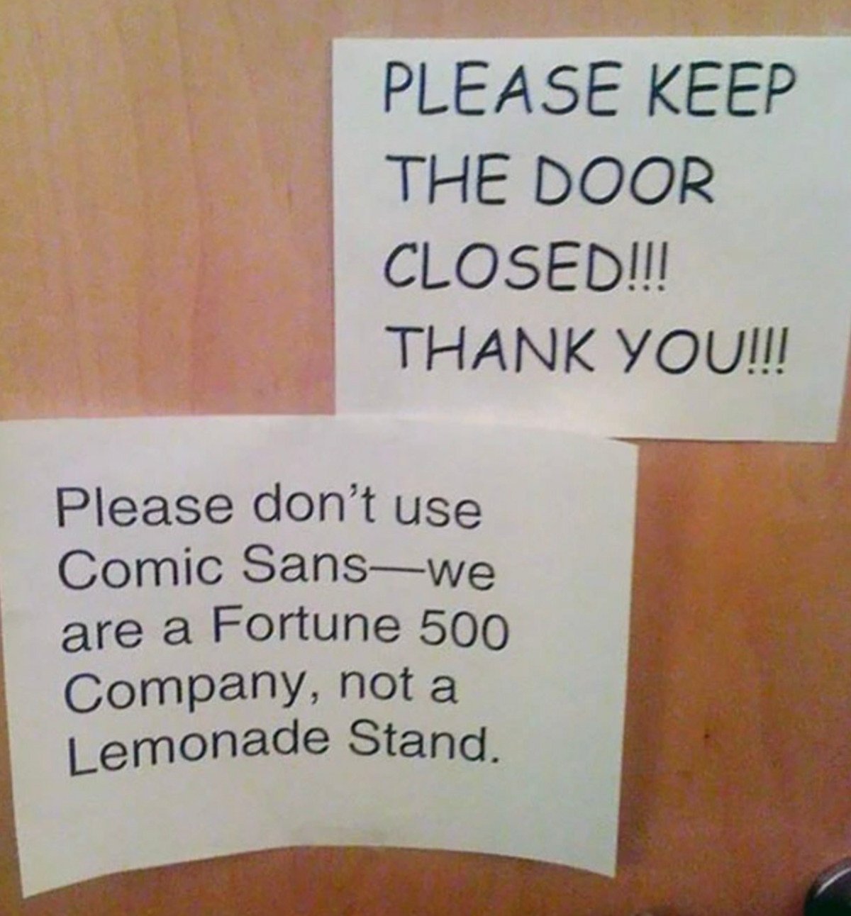 Two printed signs on a wooden door. The top sign asks to keep the door closed. The bottom sign humorously requests not to use Comic Sans font, stating they are a Fortune 500 company, not a lemonade stand.