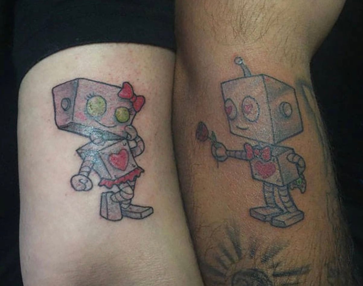 Two forearms with matching robot tattoos: one is a female robot with a bow and heart, the other is a male robot holding a rose, both with boxy designs and affectionate expressions.
