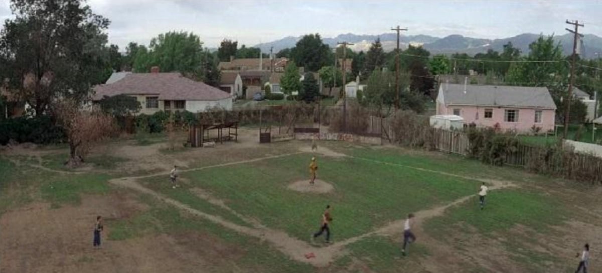 A group of kids play baseball on a grassy, makeshift field surrounded by wooden fences, with houses and trees in the background under a cloudy sky.