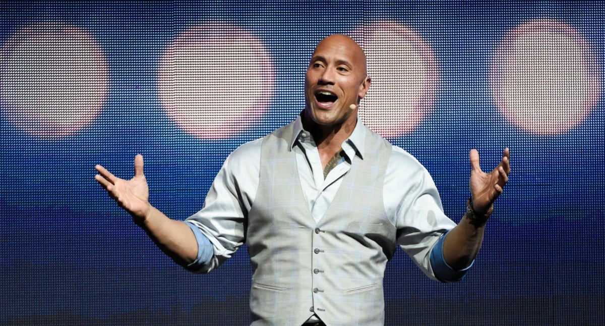 A smiling man wearing a light-colored vest and shirt speaks on stage with arms open wide, standing in front of a background of large, blurred circular lights.