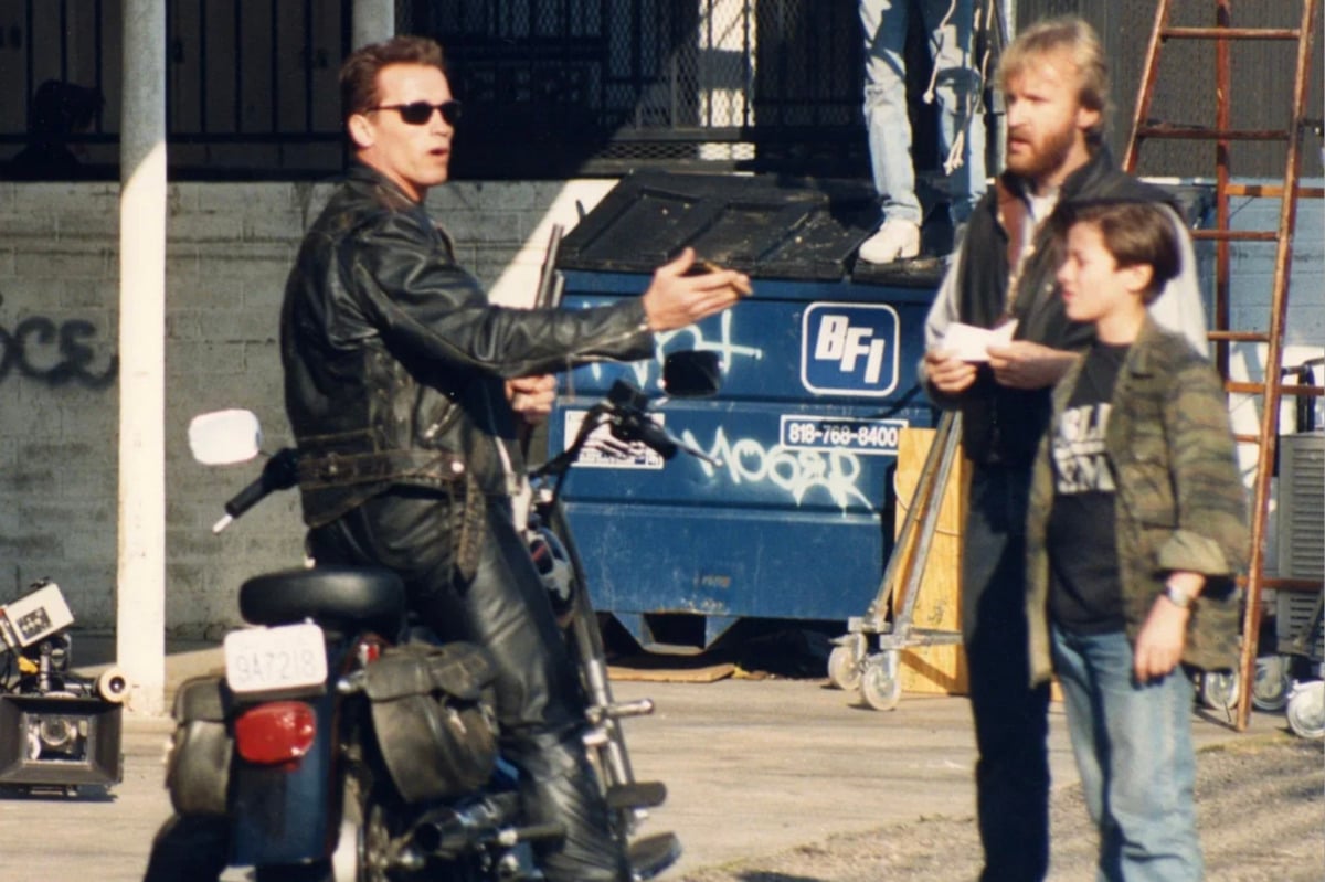 A man in black leather sits on a motorcycle, gesturing with his hand toward two people standing nearby—one adult holding papers and one child. They are outside near a blue dumpster and film equipment.