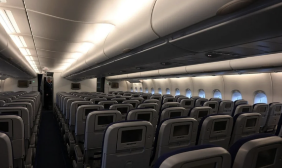 Empty airplane cabin with rows of gray seats, each equipped with a personal entertainment screen. Overhead compartments are closed, and natural light comes in through the windows on the right side.