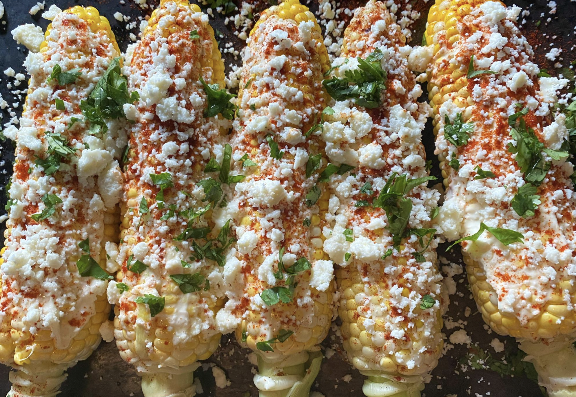 Five ears of Mexican street corn (elote) topped with crumbled cheese, chopped cilantro, and chili powder are arranged in a row on a dark surface.