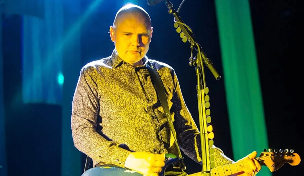 A bald man wearing a patterned shirt plays an electric guitar under bright stage lights, standing in front of a microphone during a live music performance.