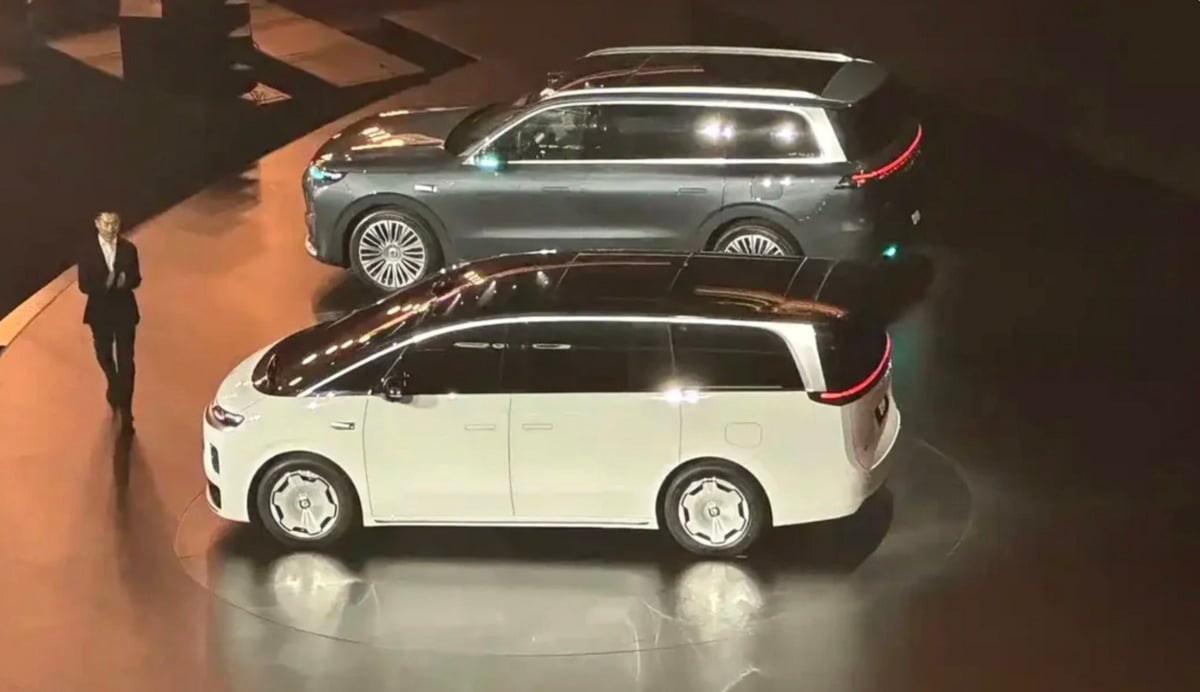Two electric vehicles, a white minivan and a grey SUV, are displayed on a stage with a person standing nearby under bright lights. The vehicles feature modern, sleek designs with prominent rear light bars.