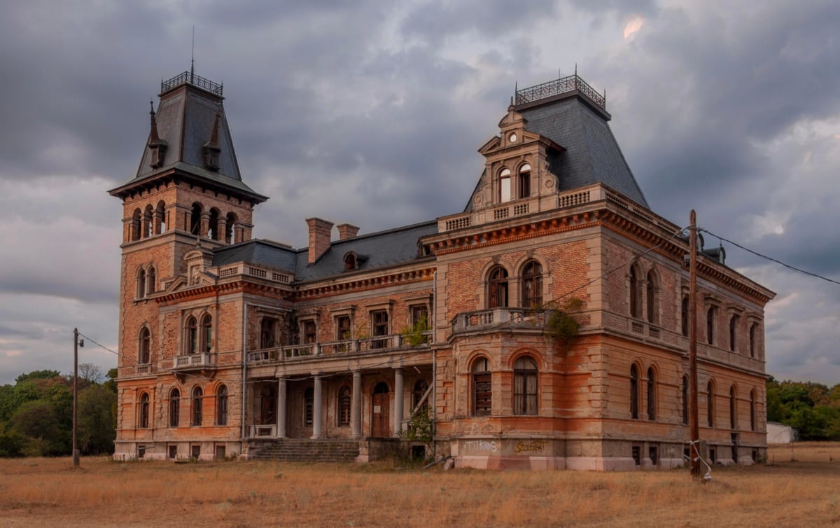 An old, abandoned mansion with ornate architecture, tall towers, and boarded windows stands in a dry, grassy field under a cloudy sky. Some vegetation grows on parts of the building, giving it a decayed, eerie appearance.