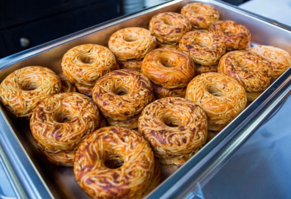 A metal tray filled with round, bagel-shaped foods made from baked spaghetti, with a brown and golden crust, arranged closely together.