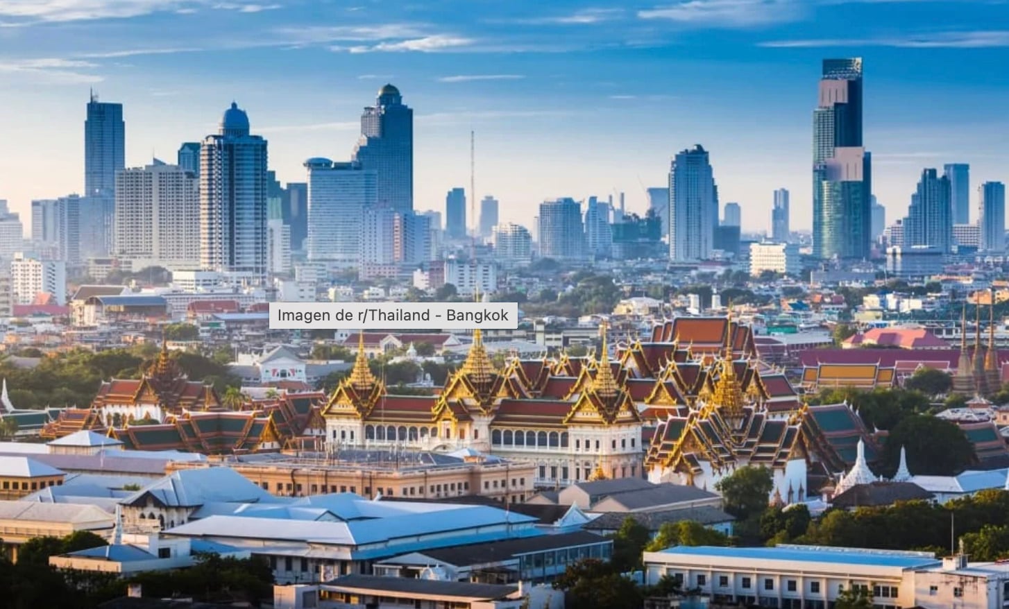 Modern skyscrapers rise behind the ornate golden rooftops of traditional Thai temples in central Bangkok, Thailand, under a blue sky with scattered clouds.