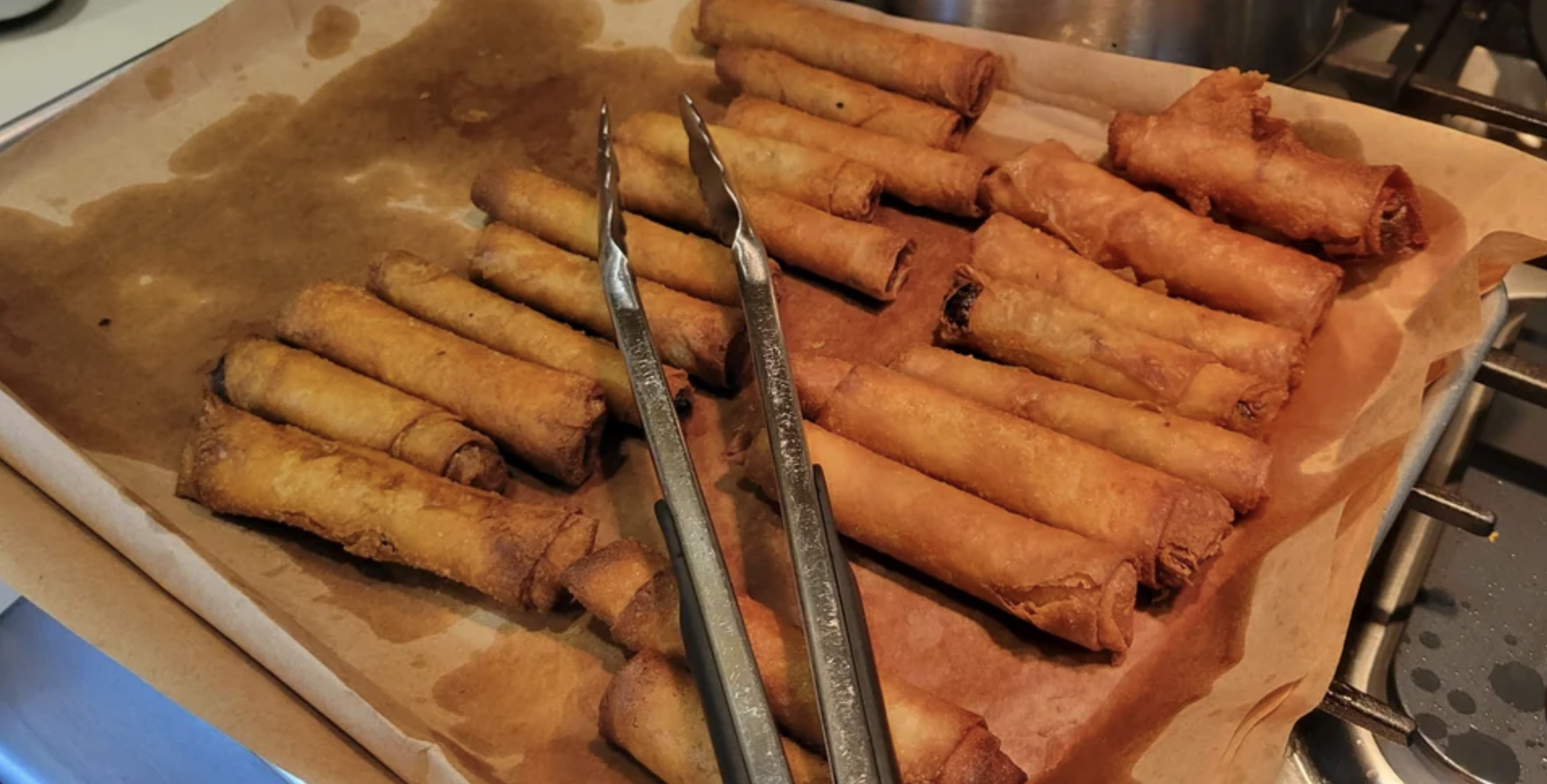 A baking tray lined with parchment paper holds several golden-brown fried spring rolls, with a pair of metal tongs resting on top, next to a stove.