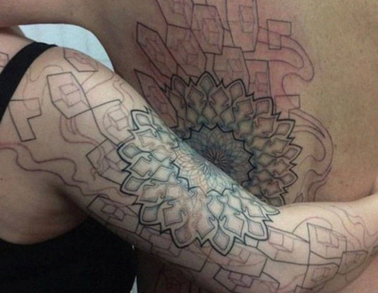 Two people with black ink geometric tattoos on their arm and back, featuring intricate, overlapping patterns that align perfectly when they embrace. The tattoos connect to form a larger, unified design.