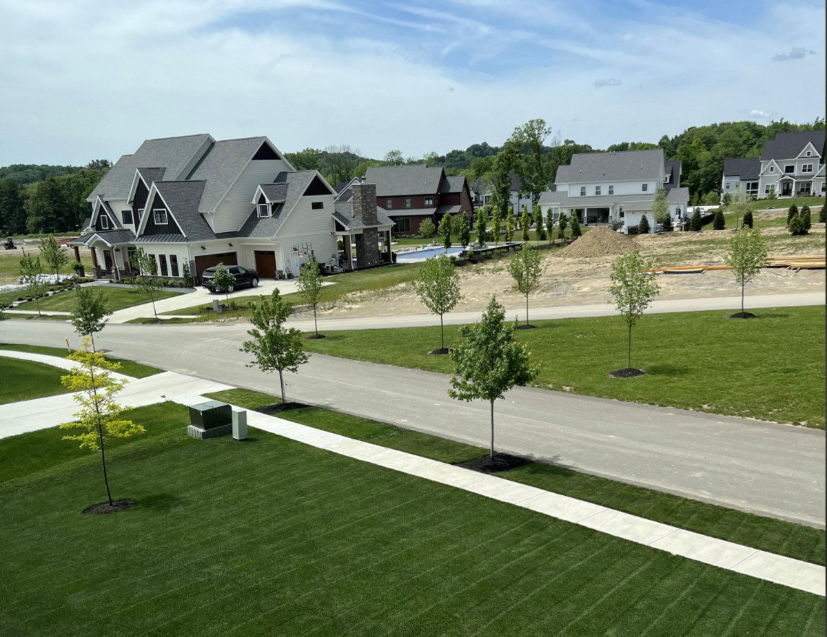 A suburban neighborhood with large houses, green lawns, young trees, and a partly cloudy sky. Some construction work is visible with dirt piles and partially built structures in the background.