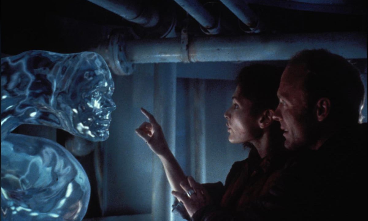 A woman reaches out to touch a large, transparent, water-like face while a man stands behind her in a dimly lit, industrial setting with pipes overhead.