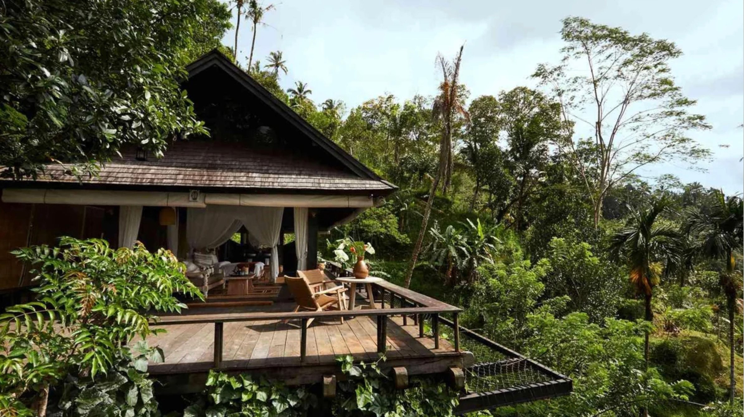 A cozy wooden cottage with an open deck and chairs overlooks lush green tropical trees and dense foliage, creating a peaceful retreat surrounded by nature.