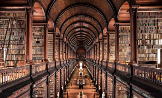 A grand, historic library hall with high, arched wooden ceilings and two stories of bookshelves filled with books, overlooking long rows of reading tables below.