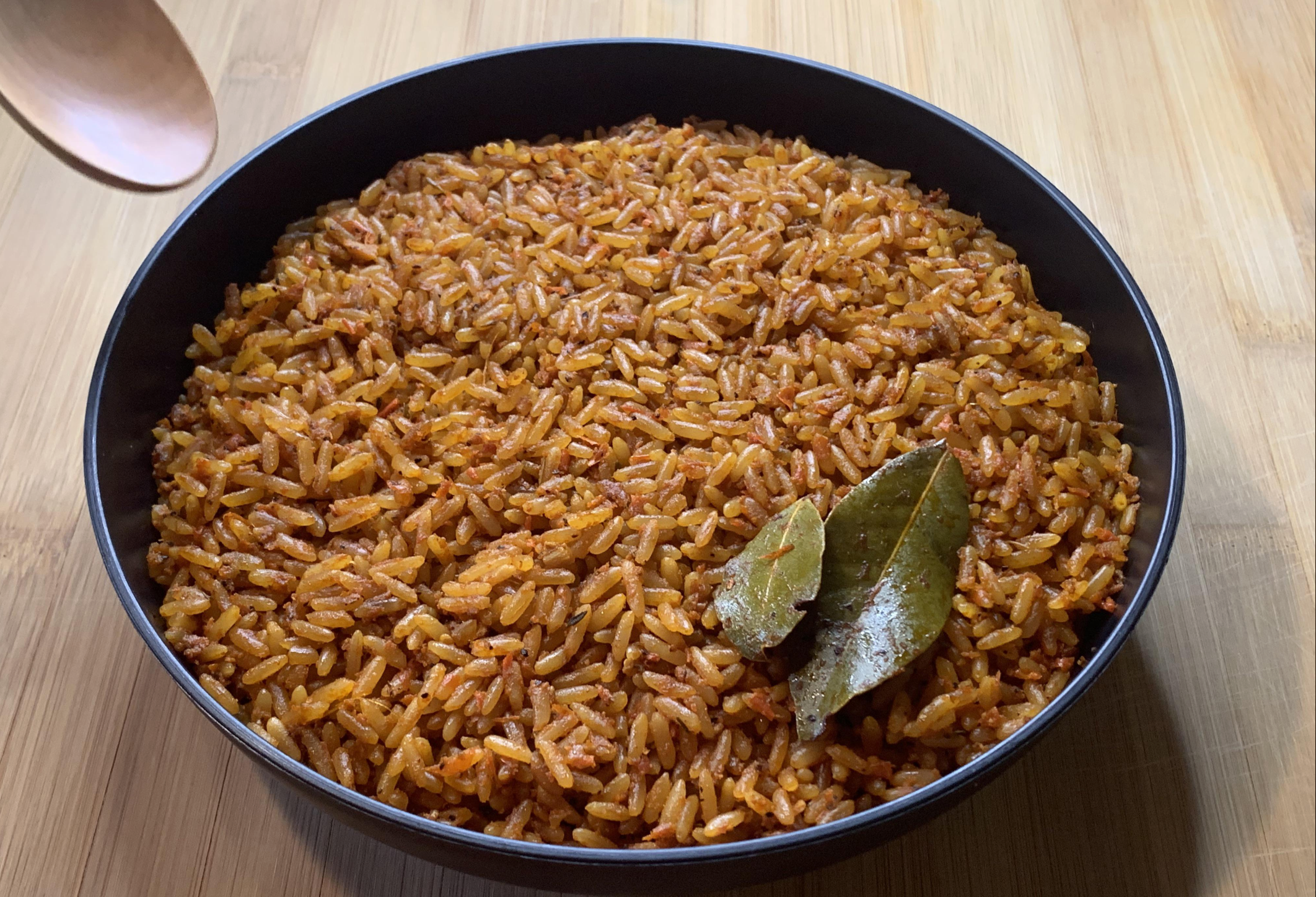A bowl of orange-red jollof rice garnished with two bay leaves, placed on a wooden surface with part of a wooden spoon visible on the left.