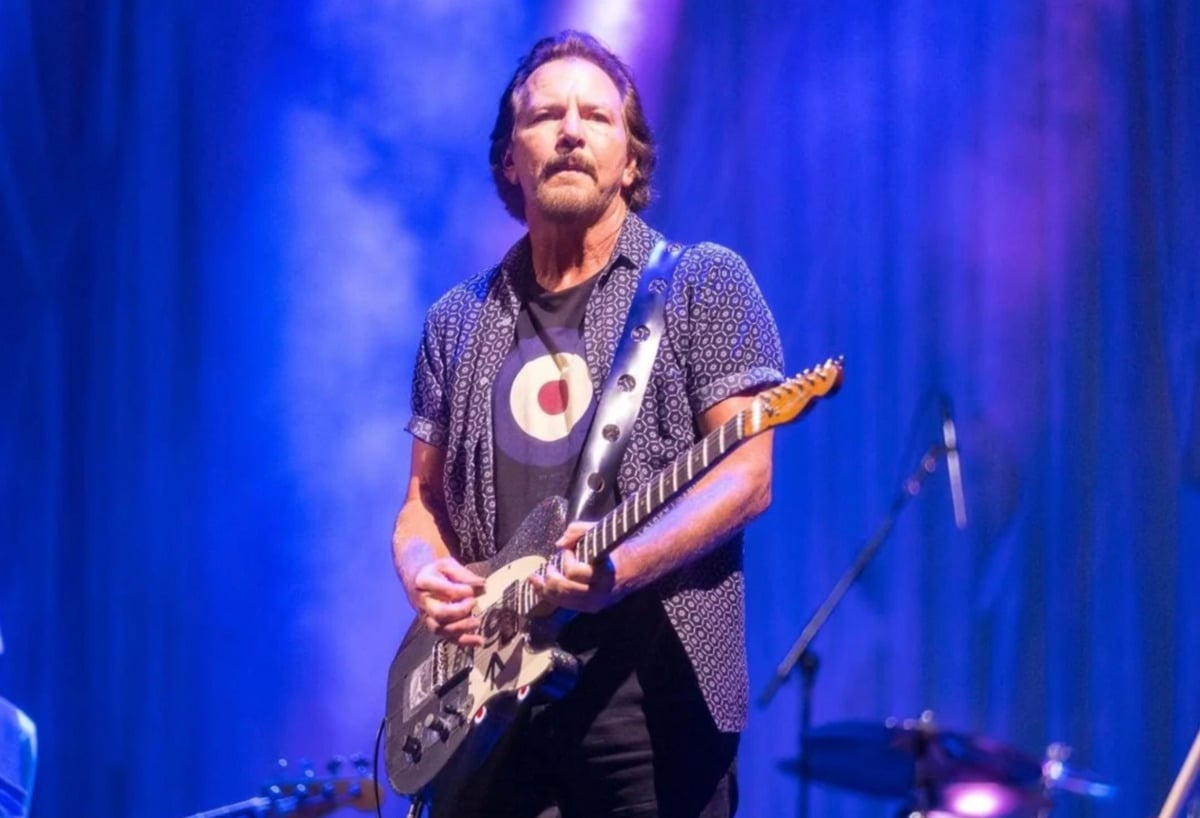 A man with a mustache and beard plays an electric guitar on stage under purple and blue lights, wearing a patterned shirt over a black t-shirt with a circular design. Drums and music equipment are visible in the background.