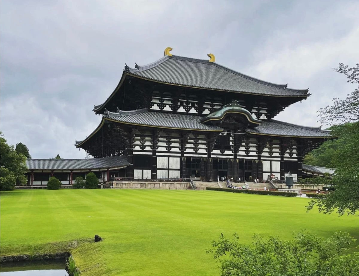 A large traditional Japanese temple with a dark tiled roof and ornate golden ornaments sits on a manicured green lawn under a cloudy sky, surrounded by trees and shrubs.