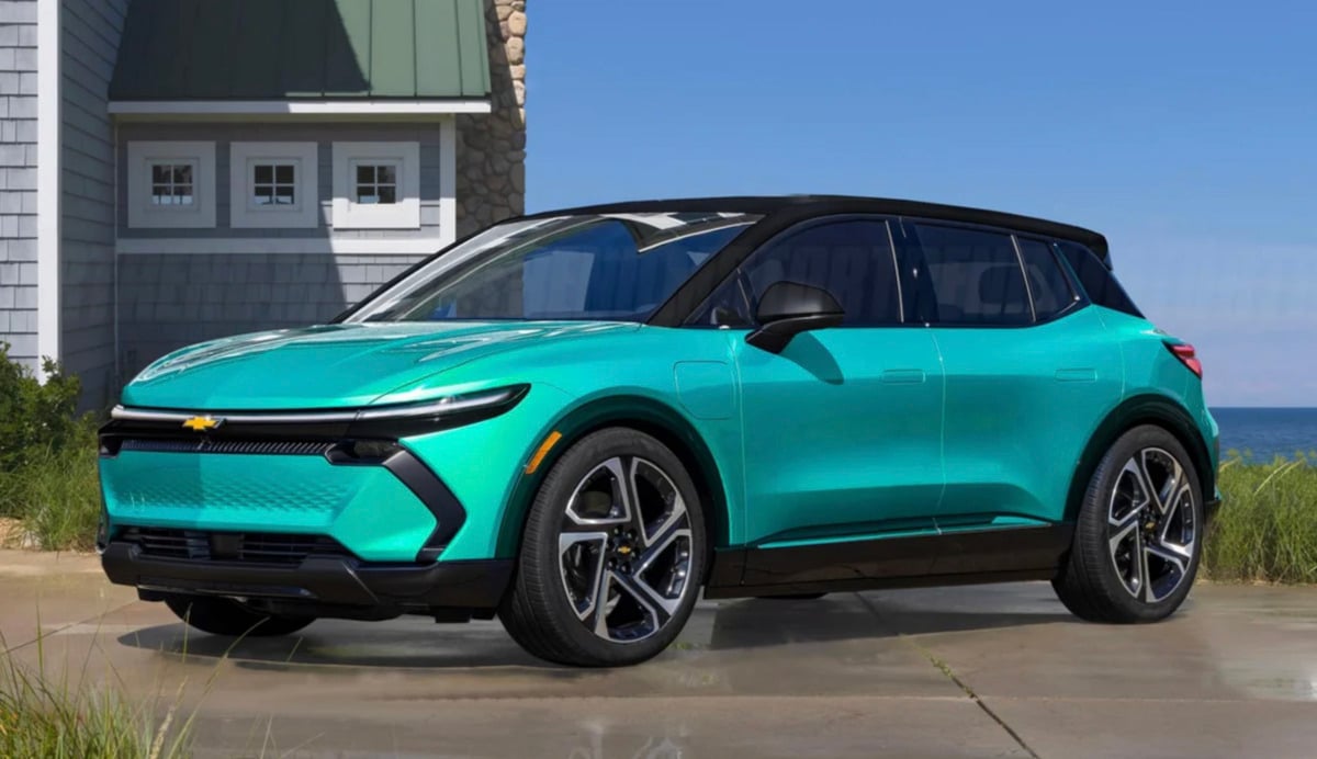 A turquoise Chevrolet electric SUV parked on a wet driveway near a beach house, with grass and the ocean visible in the background under a clear sky.