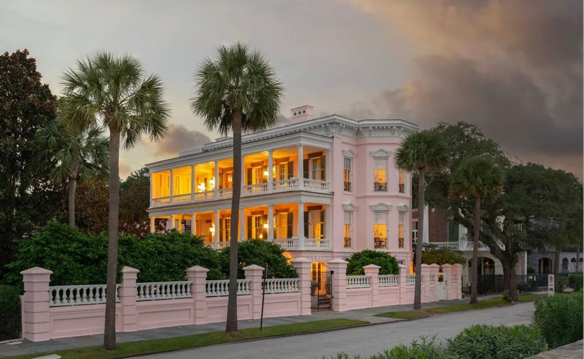 A large, elegant pink mansion with two wraparound porches, glowing warmly from inside at dusk. The home is surrounded by lush greenery, palm trees, and enclosed by a pink and white fence.