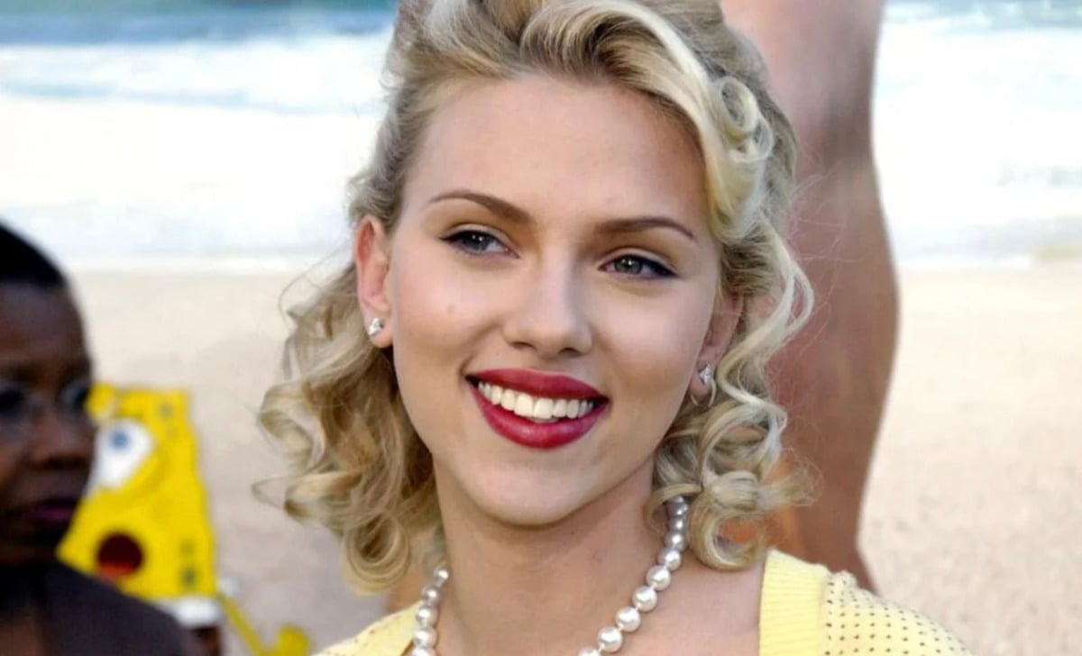 A smiling woman with curly blonde hair, red lipstick, and pearl earrings stands on a beach. She is wearing a pearl necklace and a yellow top. The background shows sand, ocean, and a blurry figure.