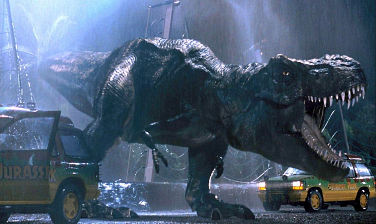 A large Tyrannosaurus rex roars in the rain, standing between two damaged Jurassic Park SUVs at night, with broken fencing and wires in the background.