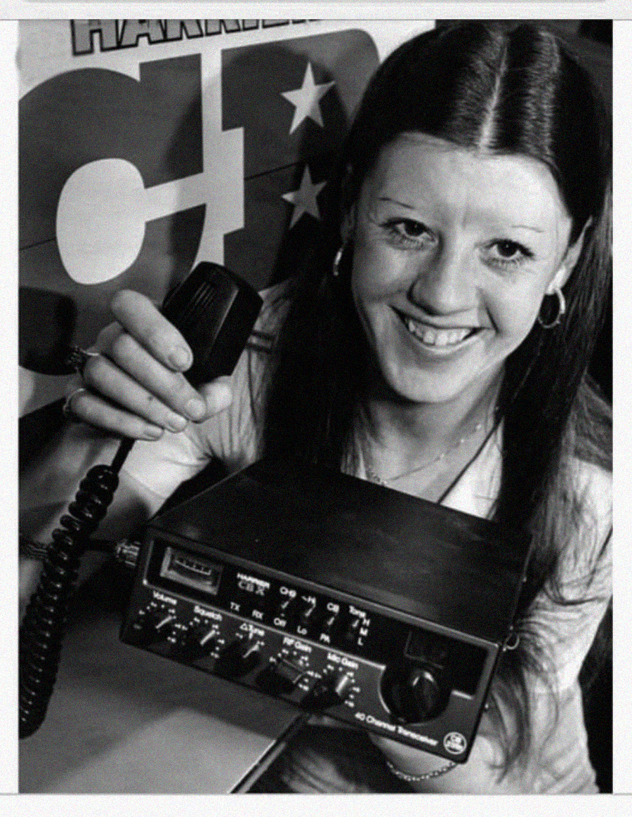 A woman smiling and holding a CB radio microphone, with a CB radio unit in front of her and a poster partially visible in the background.