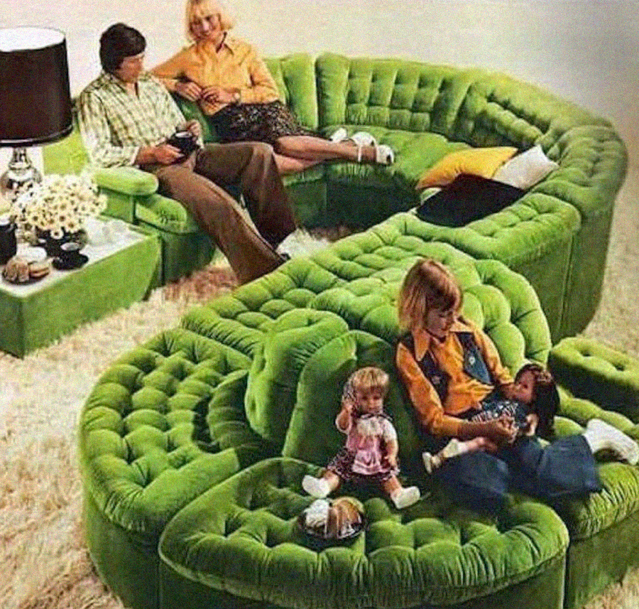A retro living room with a plush, green, curved sectional sofa. Two adults sit at the back, while a woman and two children sit at the front; the children play with dolls. The décor is vintage with a lamp and flowers on a side table.