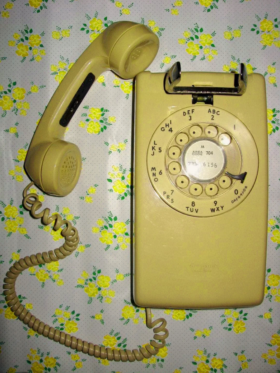 A vintage yellow rotary dial telephone with a curly cord rests on a floral-patterned surface with yellow flowers and green leaves. The receiver is placed to the side of the phone.