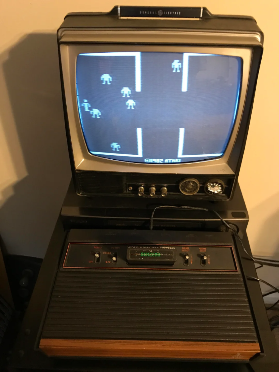 A vintage TV displays a retro video game with pixelated characters. Below, an Atari 2600 game console is connected, resting on a small table with buttons and switches visible.
