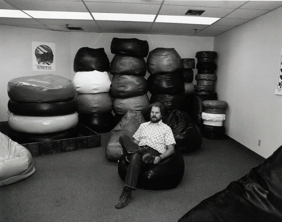 A man with a beard sits on a bean bag chair in a room filled with large stacked bean bags. The room has a carpeted floor, fluorescent lights, and posters on the walls.