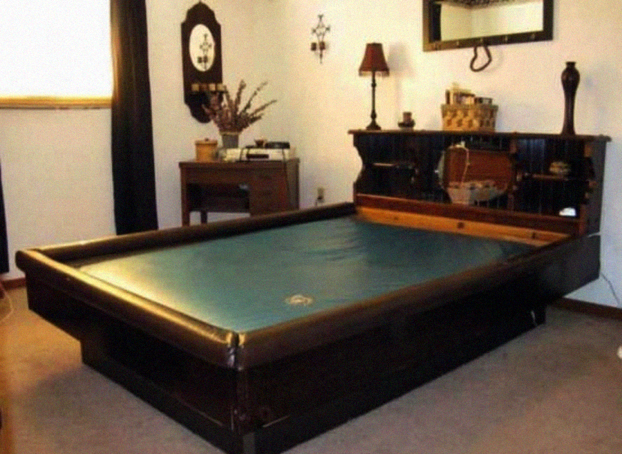 A waterbed styled to look like a pool table sits in a carpeted bedroom with vintage decor, including a mirror, lamp, and small desk in the background.