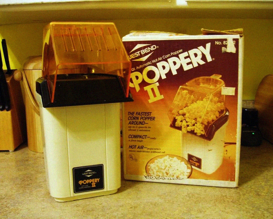 A vintage West Bend Poppery II hot air popcorn maker sits on a countertop next to its original box, which features an image of the popcorn maker and a bowl of popcorn in front.
