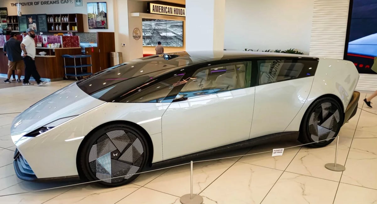 A futuristic white concept car with dark tinted windows is displayed indoors on a showroom floor, surrounded by a rope barrier. People and a coffee shop are visible in the background.