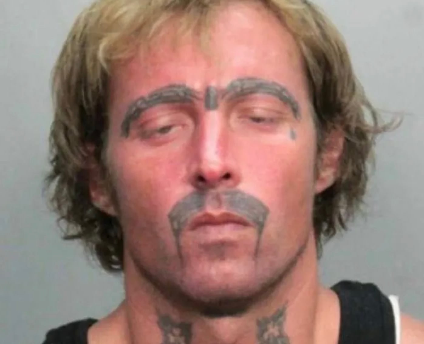 A man with messy blond hair and several facial tattoos, including tattooed eyebrows, a mustache, teardrops, and markings on his neck, has his eyes closed and is wearing a black sleeveless shirt.