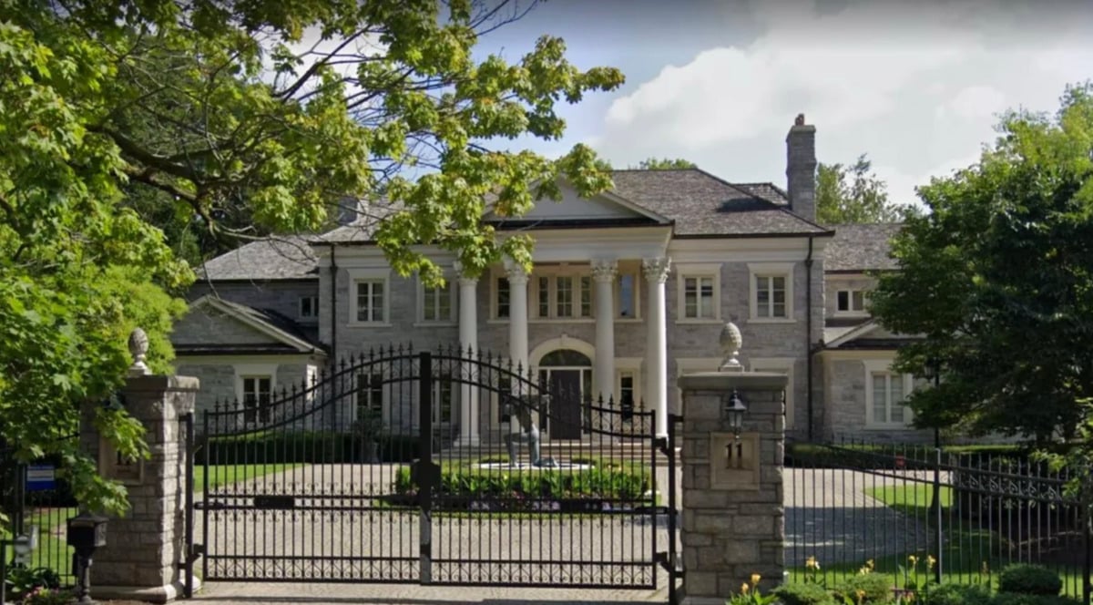 A large, elegant stone mansion with tall white columns, multiple windows, and a circular driveway sits behind a black wrought iron gate and stone pillars, surrounded by trees and greenery.
