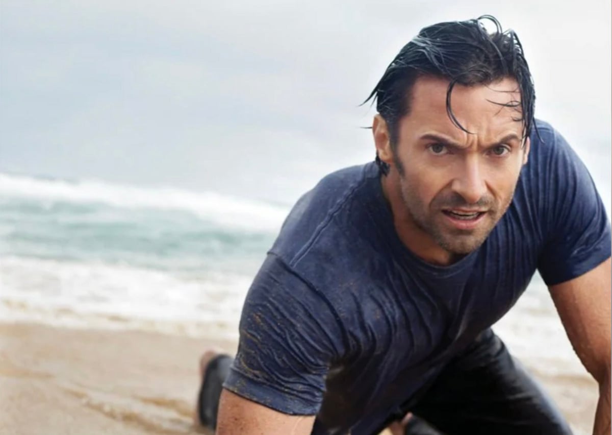 A man with wet hair and a soaked dark shirt is kneeling on a sandy beach near the ocean, looking intently ahead. The sky appears cloudy and the waves are visible in the background.