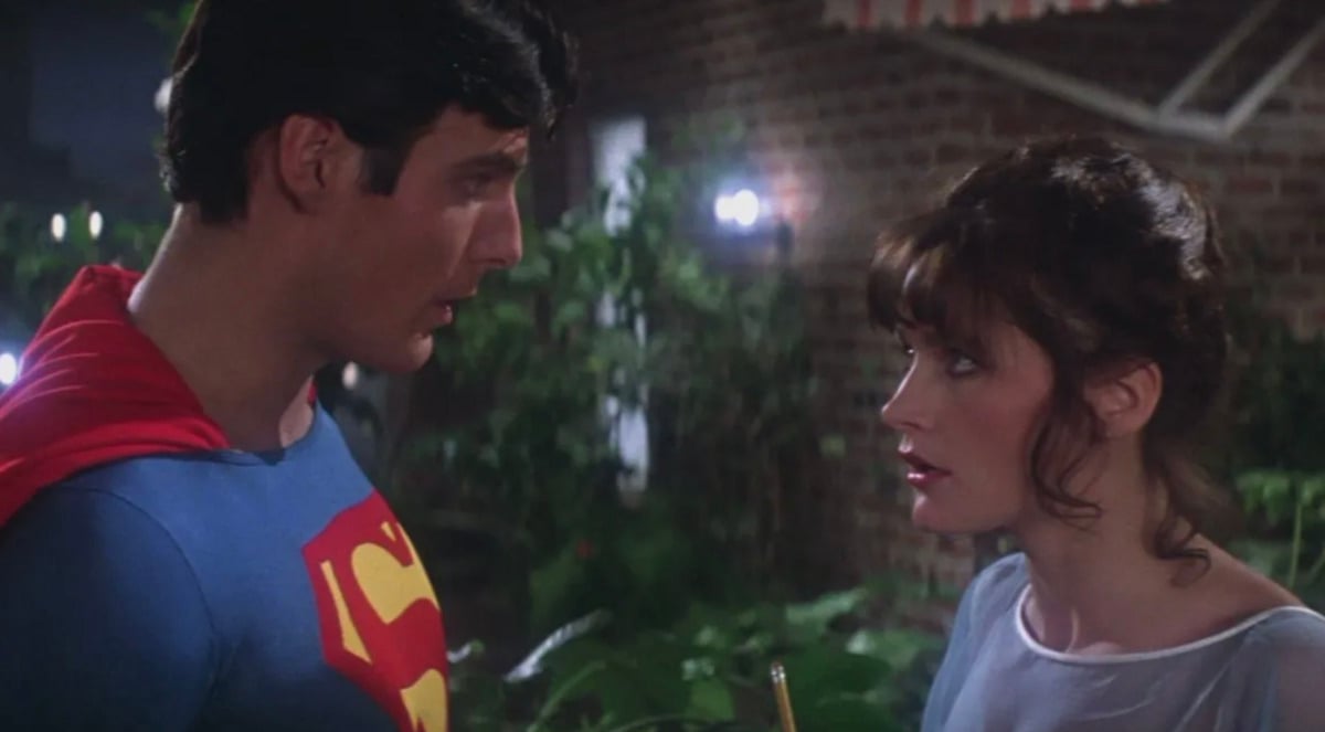 Superman, in his blue suit and red cape, stands facing a woman with brown hair and a white top, outdoors at night with greenery and brick walls in the background. They appear to be having a serious conversation.