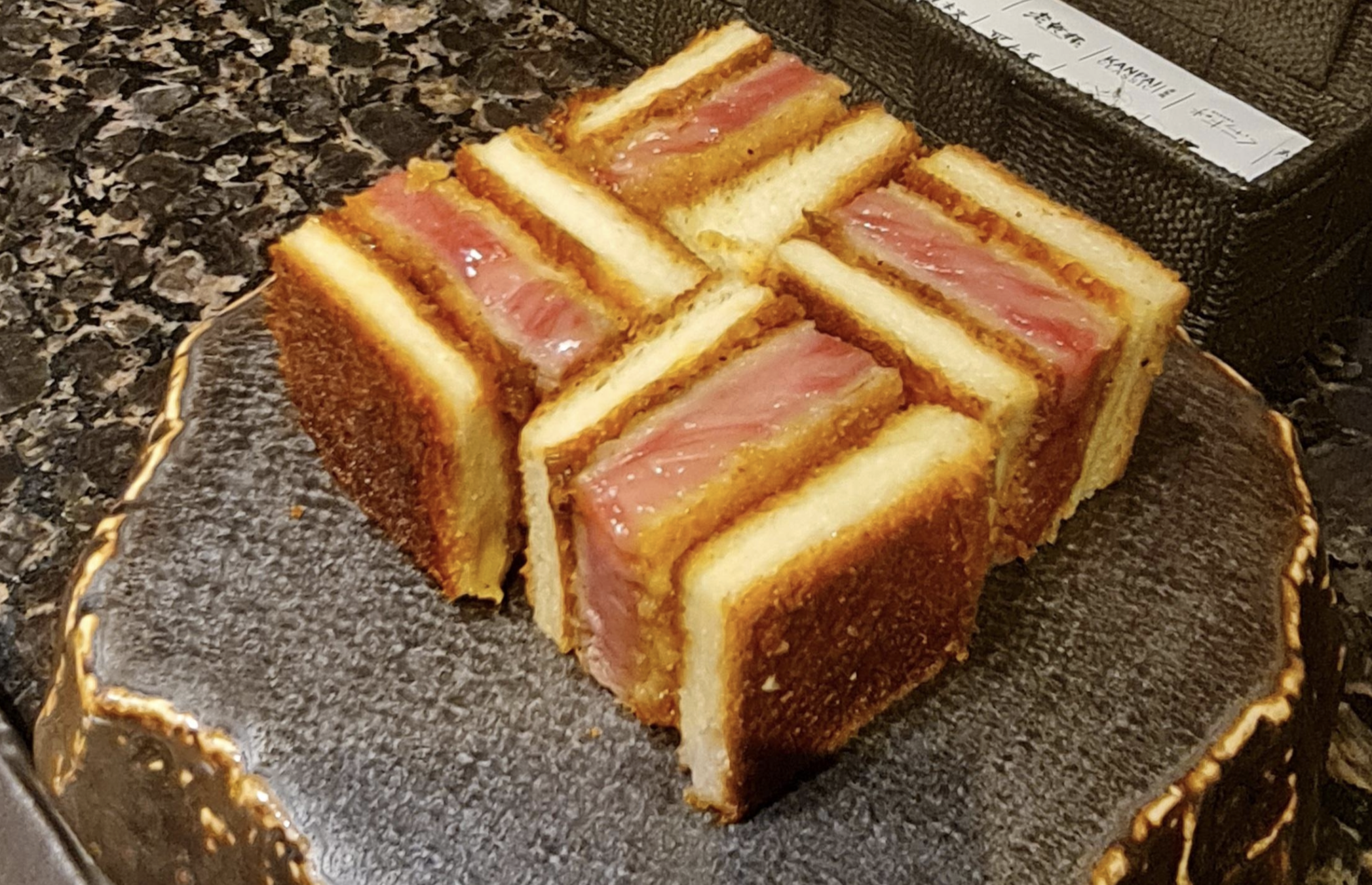 Six pieces of wagyu katsu sando, featuring thick slices of breaded, fried steak between toasted bread, are arranged neatly on a decorative stone plate.
