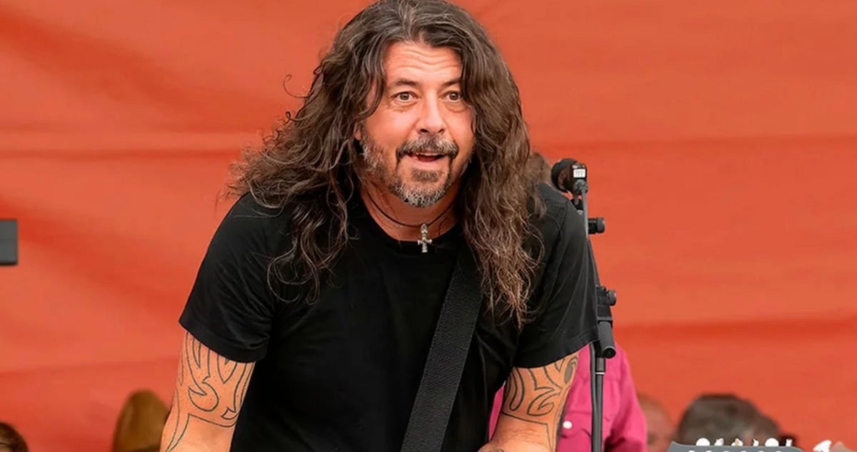 A man with long brown hair and a beard, wearing a black t-shirt and displaying arm tattoos, plays guitar and smiles on stage in front of an orange backdrop.