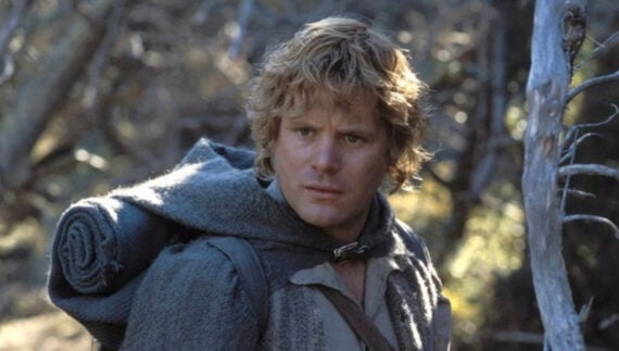 A person with curly blond hair wearing a gray cloak and carrying a rolled blanket on their back stands in a wooded area, looking slightly to the side with a serious expression.