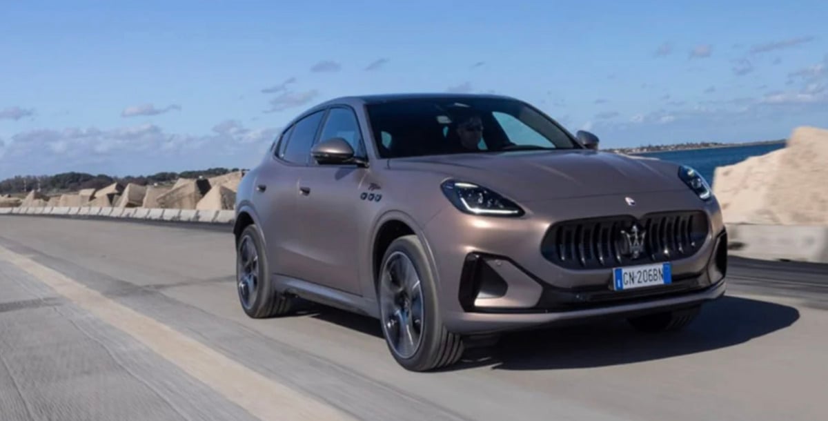 A brown Maserati SUV drives on a coastal road with stone barriers and blue sky in the background.