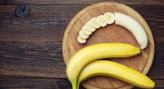 Two whole bananas and a half-peeled banana, partially sliced, lie on a round wooden cutting board atop a dark wooden surface.