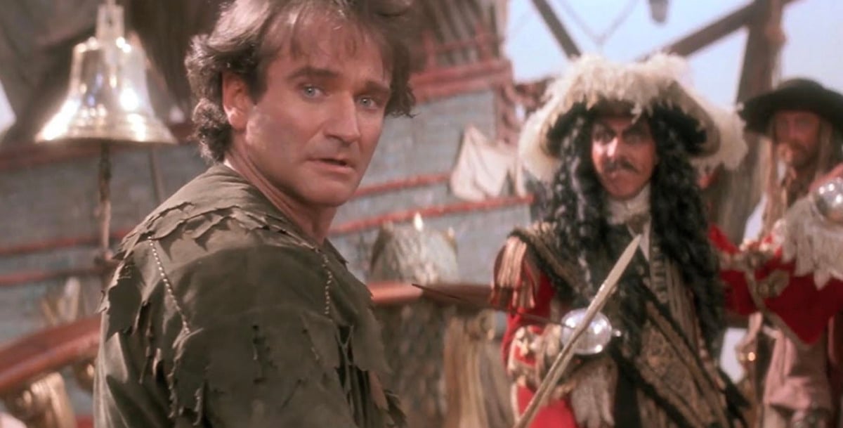 A man in tattered clothes stands on a ship deck, looking worried, while a man in an elaborate red coat and feathered hat wields a sword behind him. Other pirates are visible in the background.