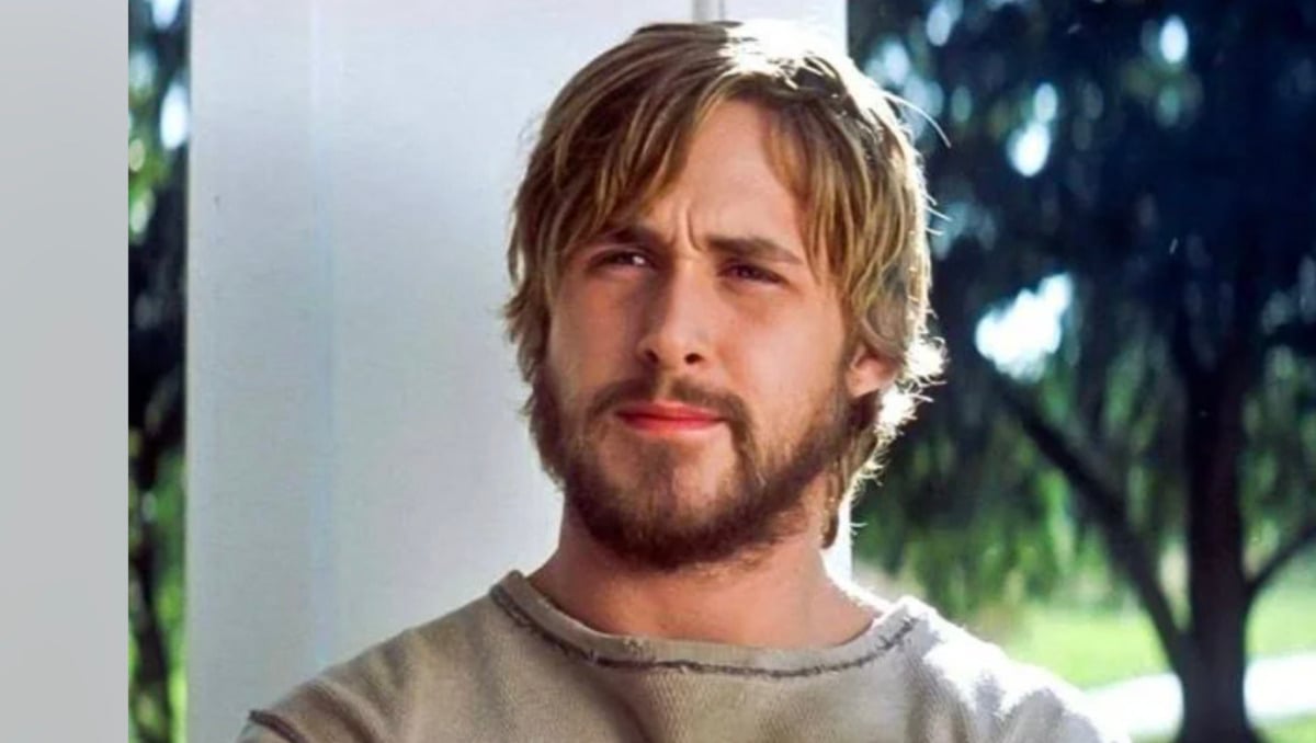 A man with light brown hair and a beard stands outside in natural light, wearing a beige sweater. He looks slightly to the side with a thoughtful expression, with trees and greenery blurred in the background.